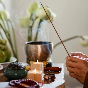 May include: A wooden table with a green ceramic incense burner, two lit candles, a copper incense holder, and a wooden bowl. A person's hand is holding a lit incense stick with smoke rising from it.