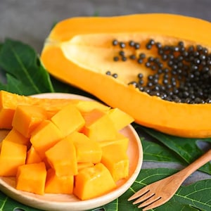May include: A ripe papaya cut into cubes on a wooden plate, with a whole papaya in the background. The papaya is a bright orange color and the seeds are black.