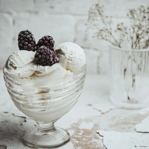 May include: A clear glass bowl filled with white vanilla ice cream topped with three frozen blackberries. The bowl is sitting on a white textured surface.