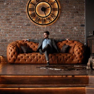 May include: A man sits on a brown leather tufted sofa with a large clock with Roman numerals on the wall behind him.