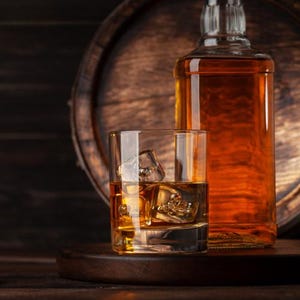 May include: A glass of whiskey with ice cubes sits on a wooden tray, next to a full bottle of amber-colored liquor. The background features a wooden barrel, creating a rustic and warm atmosphere. The image highlights the drink and its presentation.