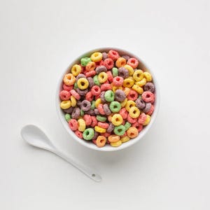 May include: A white bowl filled with colorful cereal rings. The cereal is a mix of red, yellow, green, and purple. A white spoon is next to the bowl.