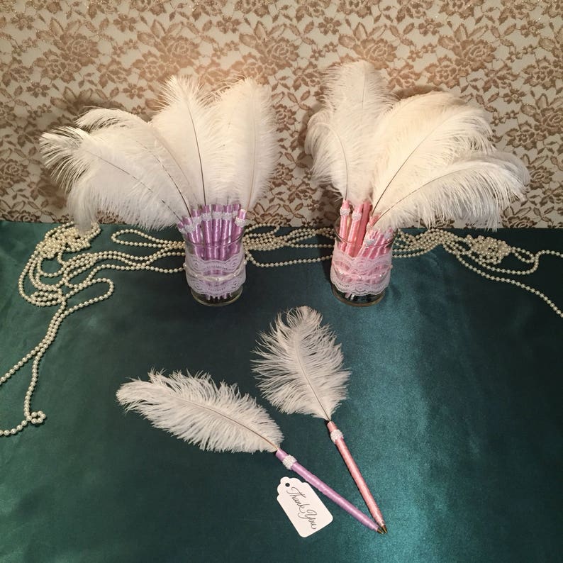 May include: Two glass vases filled with white ostrich feathers, each vase contains pink and purple pens with a white lace trim. Two white ostrich feather pens with pink and purple barrels are on a green surface with a white tag that says "Thank You".
