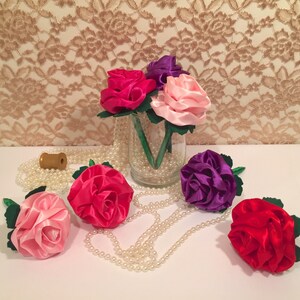 May include: A bouquet of five silk roses in pink, red, purple, and fuchsia colors. The roses are arranged in a clear glass vase with pearl beads. The roses are on a white surface with a string of pearls.