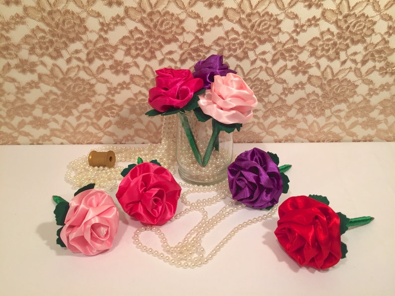 May include: A bouquet of five silk roses in pink, red, purple, and fuchsia colors. The roses are arranged in a clear glass vase with pearl beads. The roses are on a white surface with a string of pearls.