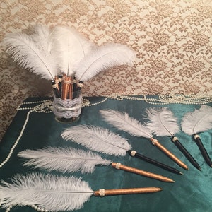 May include: A set of white feather pens with gold and black accents. The pens are arranged in a vase with a lace trim. The pens are on a green surface with a pearl necklace.