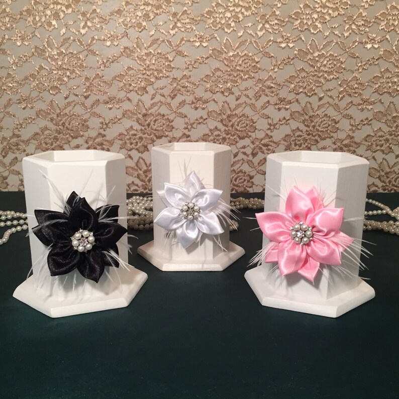 May include: Three white hexagonal pencil holders with decorative flowers. The flowers are made of white, black, and pink satin ribbon with white feathers and a rhinestone center.