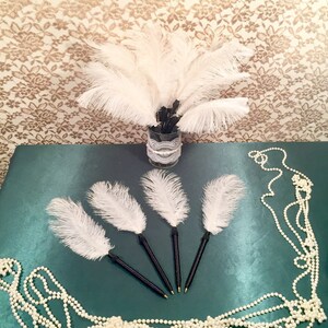May include: Five white feather pens with black handles. The pens are arranged in a star shape on a green surface. A string of white pearls is in the background.