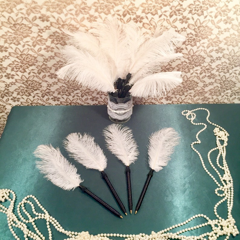 May include: Five white feather pens with black handles. The pens are arranged in a star shape on a green surface. A string of white pearls is in the background.