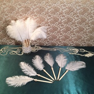 May include: White ostrich feather pens with pearl accents. The pens are arranged in a fan shape on a teal surface.