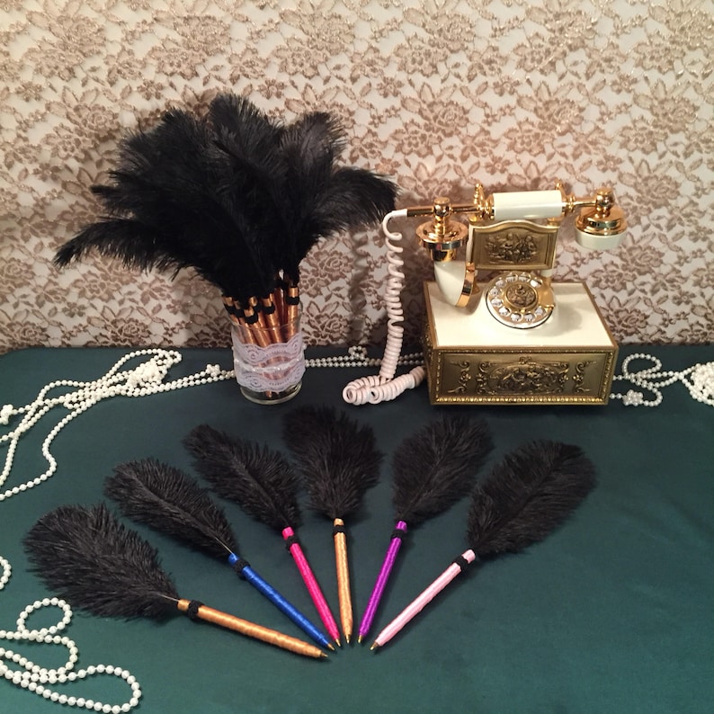 May include: A set of seven black feather pens with gold colored barrels. The pens are arranged on a green surface with a vintage rotary phone and a vase of black feathers in the background.