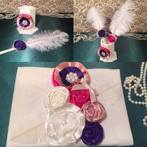 May include: A white satin pillow with a pink, white, and purple flower arrangement. The flowers are made of satin ribbon and have pearl accents. Two white feathers are on the pillow.
