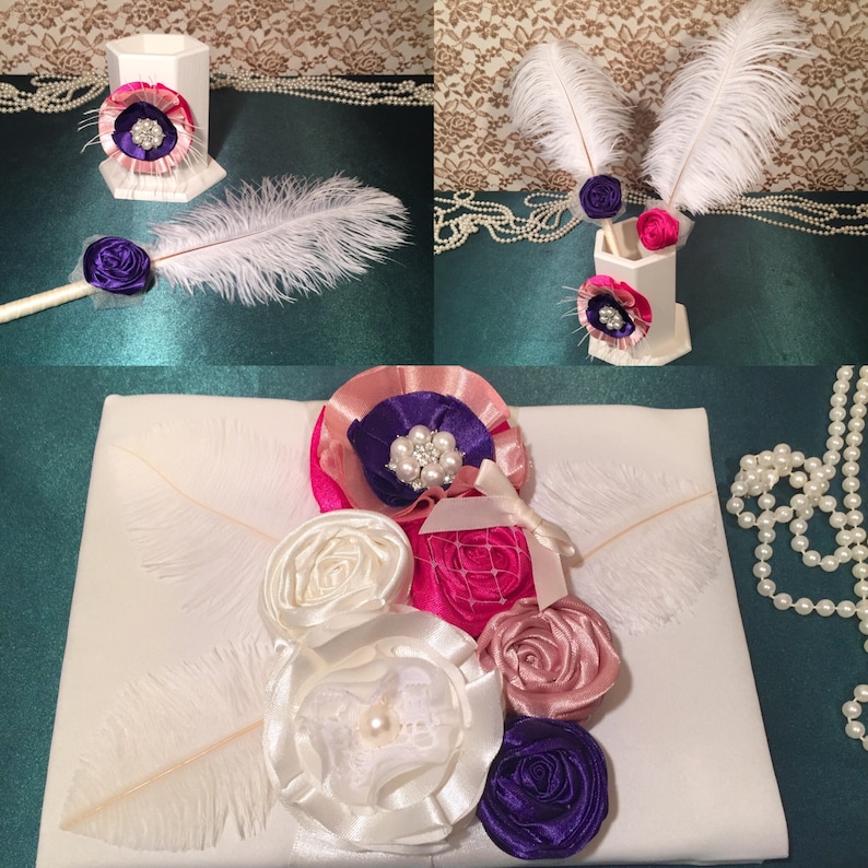 May include: A white satin pillow with a pink, white, and purple flower arrangement. The flowers are made of satin ribbon and have pearl accents. Two white feathers are on the pillow.