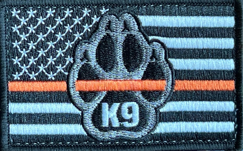 May include: Black and white embroidered patch with an American flag design and a paw print with the text "K9" in the center. A thin orange stripe is across the flag.