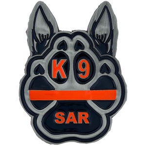 May include: A K9 SAR badge in the shape of a paw print and dog head. The badge is primarily gray, black, and orange. The text "K 9" and "SAR" are in orange. An orange line runs horizontally across the paw print.