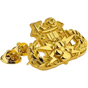 May include: Gold-colored metal K9 badge with a paw print, lightning bolts, and a shield with the letters K9. The badge is surrounded by a laurel wreath and has a pin back.