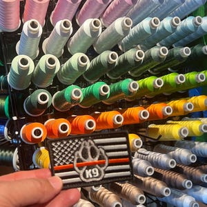 May include: A black and white American flag patch with a red paw print and the text "K9" in the center. The patch is displayed in front of a wall of colorful spools of thread.