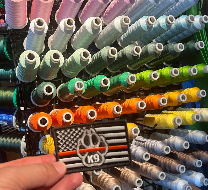 May include: A black and white American flag patch with a red paw print and the text "K9" in the center. The patch is displayed in front of a wall of colorful spools of thread.