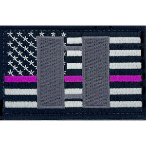 Puede incluir: Un parche bordado con un diseño de bandera estadounidense en blanco y negro. La bandera tiene una fina franja morada en la parte superior y dos torres grises en el centro. Las torres representan las Torres Gemelas del World Trade Center.