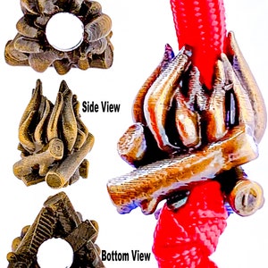 May include: A bronze-coloured campfire charm with a triangular shape. The charm is designed to look like a pile of logs with flames rising from the top. The charm is shown on a red cord.
