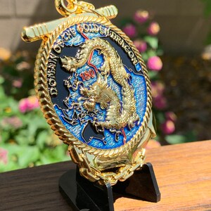 May include: A gold-colored coin with a detailed dragon design, the text "DOMAIN OF THE GOLDEN DRAGON", and the number "180". The coin is displayed on a black stand, with a blue and black background.