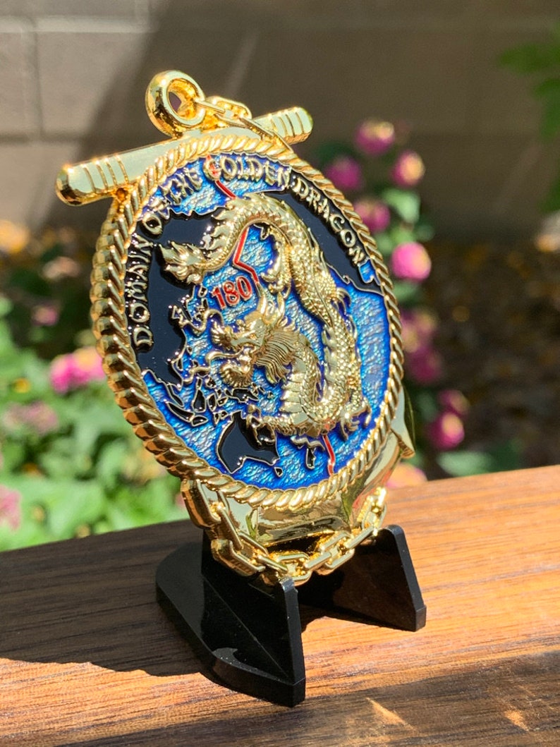 May include: A gold-colored coin with a detailed dragon design, the text "DOMAIN OF THE GOLDEN DRAGON", and the number "180". The coin is displayed on a black stand, with a blue and black background.