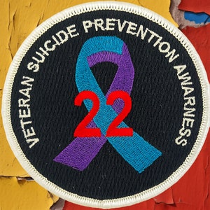 Könnte beinhalten: Ein schwarzer und weißer runder Patch mit weißem Rand. Der Patch zeigt ein lila und blaues Band mit der Zahl 22 in Rot. Der Text "Veteran Suicide Prevention Awareness" ist um den Rand des Patches geschrieben.