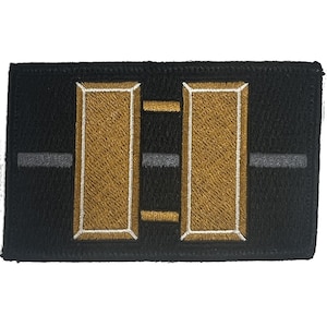 May include: A rectangular black embroidered patch featuring two vertical gold bars outlined in white, connected by two horizontal gold bars, with a thin gray horizontal line running through the center of the entire design.