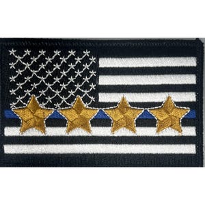 May include: Embroidered patch featuring a stylized American flag design. The flag has a black and white striped background with a blue line and four gold stars. The stars are embroidered with gold thread and outlined in white.