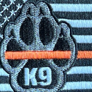May include: A black and white paw print with the text "K9" in white. The paw print is on a thin orange stripe, which is on top of a black and white American flag.