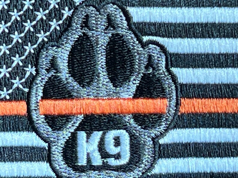 May include: A black and white paw print with the text "K9" in white. The paw print is on a thin orange stripe, which is on top of a black and white American flag.