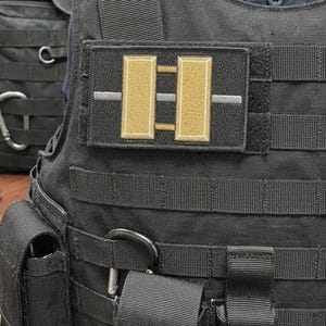 以下が含まれることがあります： 黒いタクティカルベスト。中央には、細い銀色の水平線で区切られた2本の垂直な金色のバーが描かれた長方形のパッチが付いています。ベストには複数の水平ウェビングストラップ、統合されたポケット、透明なプラスチック製のIDカードホルダーが備わっています。