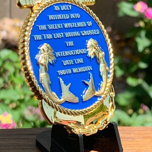 May include: A gold-colored, oval-shaped medallion with a blue center, featuring two mermaid figures and text. The text reads "AS DULY INITIATED INTO THE SILENT MYSTERIES OF THE FAR EAST HAVING CROSSED THE INTERNATIONAL DATE LINE 180TH MERIDIAN."