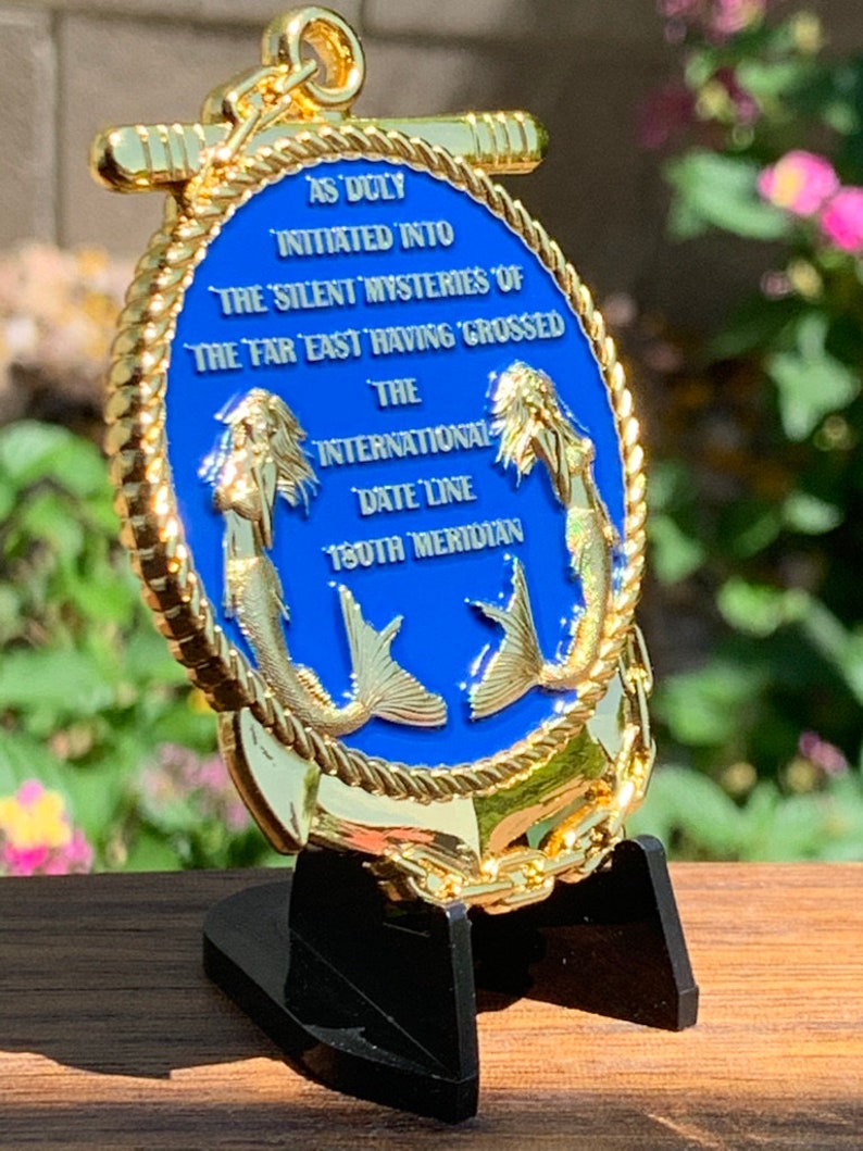 May include: A gold-colored, oval-shaped medallion with a blue center, featuring two mermaid figures and text. The text reads "AS DULY INITIATED INTO THE SILENT MYSTERIES OF THE FAR EAST HAVING CROSSED THE INTERNATIONAL DATE LINE 180TH MERIDIAN."