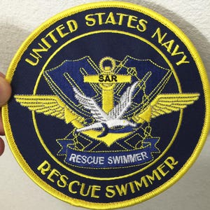 May include: A circular navy blue and yellow patch with the text "UNITED STATES NAVY" and "RESCUE SWIMMER". The patch features a central emblem with a white bird, wings, and the letters "SAR".