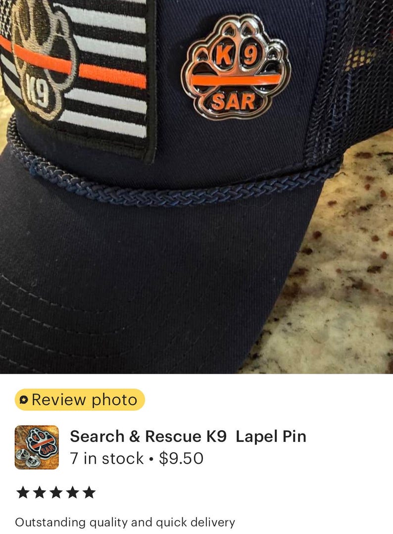 May include: A silver metal lapel pin with a paw print design. The paw print has a thin orange stripe across it and the text "K9 SAR" is inscribed on the pin.