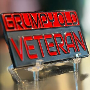 May include: A rectangular metal sign with the words "GRUMPY OLD VETERAN" in red lettering on a black and gray background. The sign is displayed on a clear plastic stand.