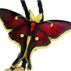 May include: A bolo tie featuring a large moth-shaped pendant with deep red and black enamel wings, gold-toned metal edges, and gold accents. The moth has a textured gold head and body, and it is attached to a black braided leather cord.