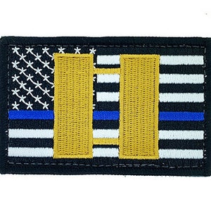 Puede incluir: Un parche de bandera estadounidense en blanco y negro con una fina línea azul, que presenta dos barras dobles doradas, que representan el rango de teniente en la aplicación de la ley.