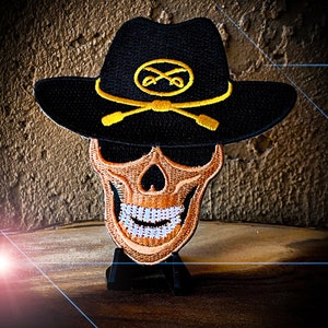 May include: A black skull wearing a black cowboy hat with a gold embroidered crossbones and a gold circle with the letters "XX" inside. The skull has a gold grin with white teeth.