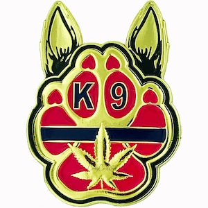 May include: Gold paw print with a black outline and red interior. The paw print has a blue and gold ribbon across the top and a green marijuana leaf in the center. The words "K9" are in the center of the paw print in blue letters.