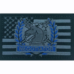 May include: A black and gray embroidered patch with an American flag background. The patch features a gray horse head with a lightning bolt and swords, surrounded by a blue laurel wreath. The text "NEGOTIATOR" is written in blue below the horse head.