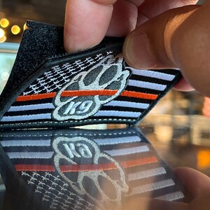 May include: A black and white embroidered patch with a paw print and the text "K9" in the center. The patch has a thin red stripe across the top and a thin white stripe across the bottom. The patch is on a black background.