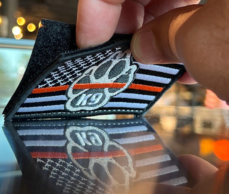 May include: A black and white embroidered patch with a paw print and the text "K9" in the center. The patch has a thin red stripe across the top and a thin white stripe across the bottom. The patch is on a black background.