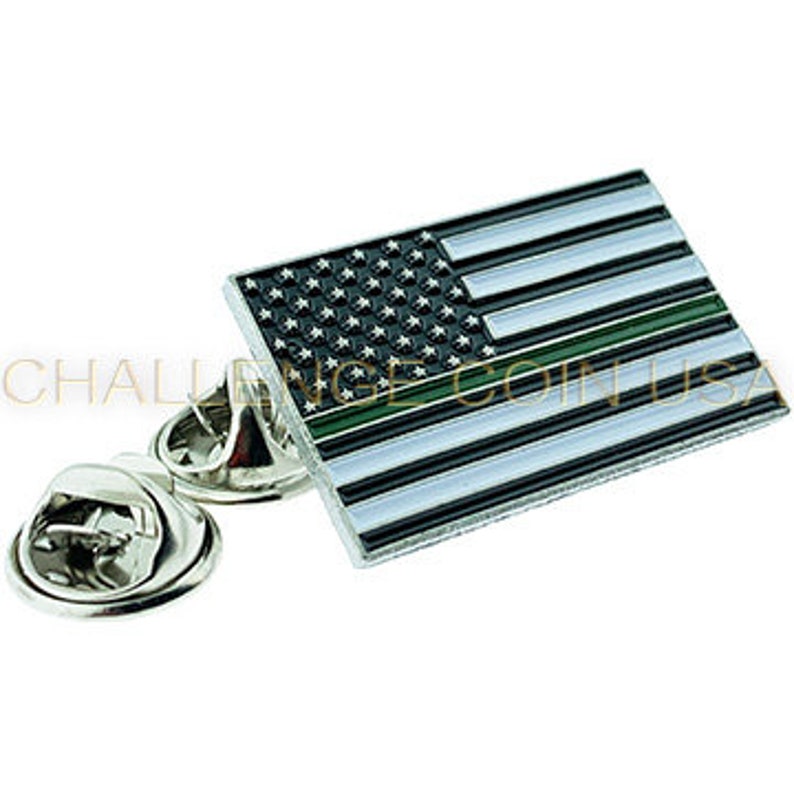 Pode incluir: Um broche de lapela prateado com uma bandeira americana de linha verde fina. A bandeira tem listras brancas, estrelas pretas e uma linha verde fina na parte inferior.