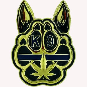 Puede incluir: Pin de esmalte dorado y negro con un diseño de huella de pata de perro. La huella de pata tiene una línea fina azul y negra en la parte superior y una hoja de marihuana verde en el centro. Las palabras "K9" están en el centro de la huella de pata.