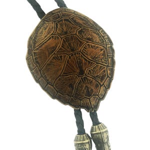 May include: A bolo tie featuring a brown, textured turtle shell centerpiece with a detailed, segmented pattern. The tie includes a black braided leather cord and two metallic silver-toned aglets at the ends, each engraved with the text True Love.