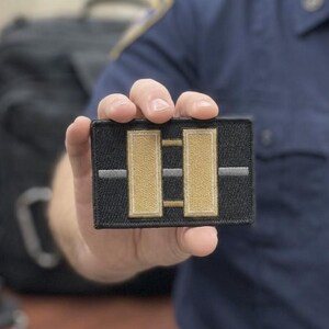 以下が含まれることがあります： 黒い刺繍が施された長方形のパッチで、中央に2本の縦長のベージュのバーが配置されています。パッチの中央には細い水平のグレーのラインが通り、両方のベージュのバーと交差しています。パッチには黒い縁取りがあり、質感のある仕上がりになっています。