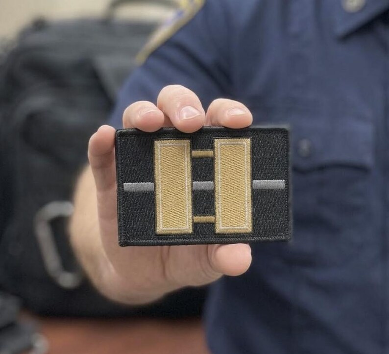 以下が含まれることがあります： 黒い刺繍が施された長方形のパッチで、中央に2本の縦長のベージュのバーが配置されています。パッチの中央には細い水平のグレーのラインが通り、両方のベージュのバーと交差しています。パッチには黒い縁取りがあり、質感のある仕上がりになっています。