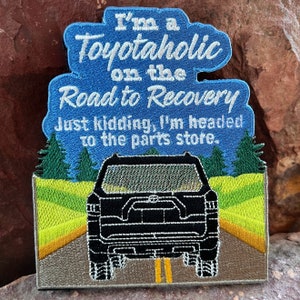 Puede incluir: Parche bordado con fondo azul y texto blanco que dice "I'm a Toyotaholic on the Road to Recovery Just kidding, I'm headed to the parts store." El parche presenta un SUV Toyota negro conduciendo por un camino con árboles verdes y hierba amarilla a cada lado.